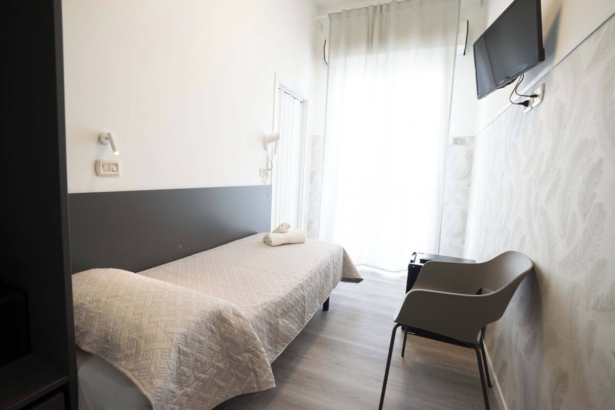 A hotel room with a bed, a chair, a TV mounted on the wall, and a window with curtains.