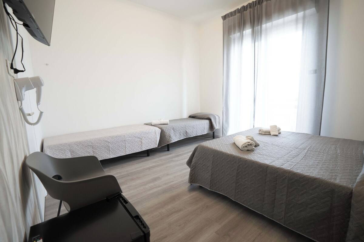 A room with two single beds, a desk, a chair, a TV, and a window with curtains.