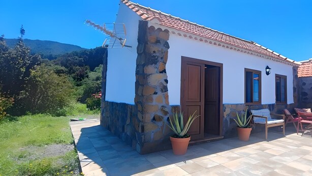 Una casa pequeña con fachada de piedra, techo de tejas y un patio con sillas y plantas en macetas.