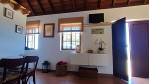 Una habitación con una mesa de comedor, sillas, un televisor, ventanas con persianas, un estante con adornos y una puerta.