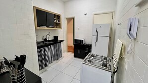 A compact kitchen with white tiled walls, a black countertop, a white refrigerator, a stove, and a microwave. - Copacabana 2 quartos na quadra da praia. (Rio de Janeiro)