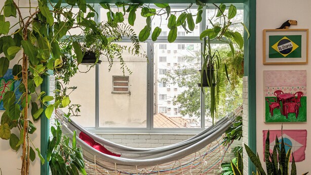 A hammock in a room with a view of buildings outside, a window with a view of buildings, and a wall with a framed picture and a plant. - Copacabana 2 quartos na quadra da praia. (Rio de Janeiro)
