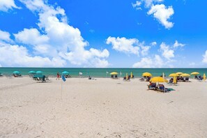 Uma praia com fileiras de espreguiçadeiras e guarda-sóis, céu claro e oceano ao fundo.