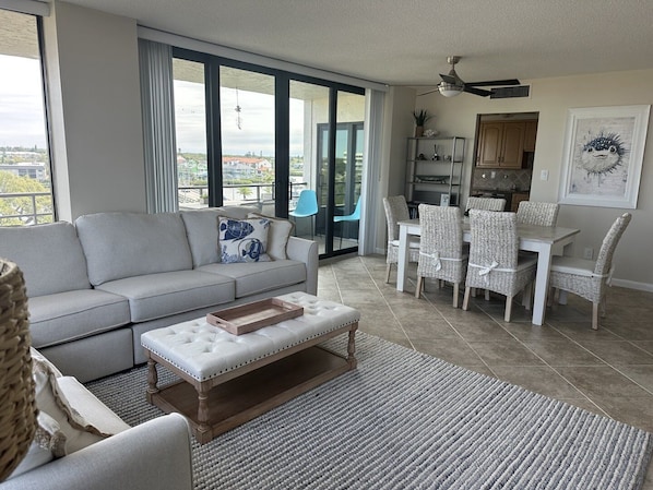 A modern living room with a sofa, ottoman, dining table with chairs, and a view through large windows.