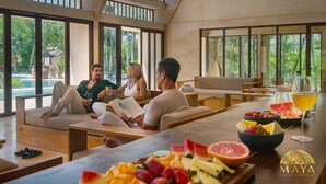 A modern lounge area with large windows, comfortable seating, and a table with fruit and drinks.