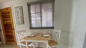 A dining area with a wooden table and white chairs, a window with blinds, and a door with a wooden panel. - Savannah Inn 204 Studio AC,Wifi,TV, (San Andrés y Providencia)