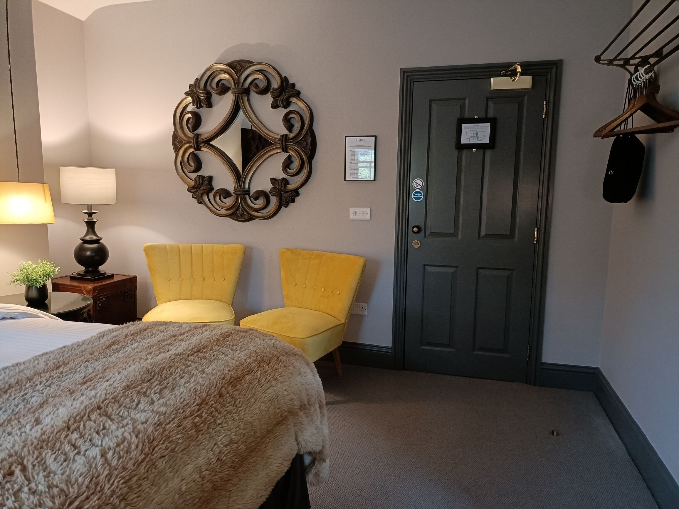 A hotel room with a bed, two yellow armchairs, a decorative wall piece, a door with a sign, and a lamp.