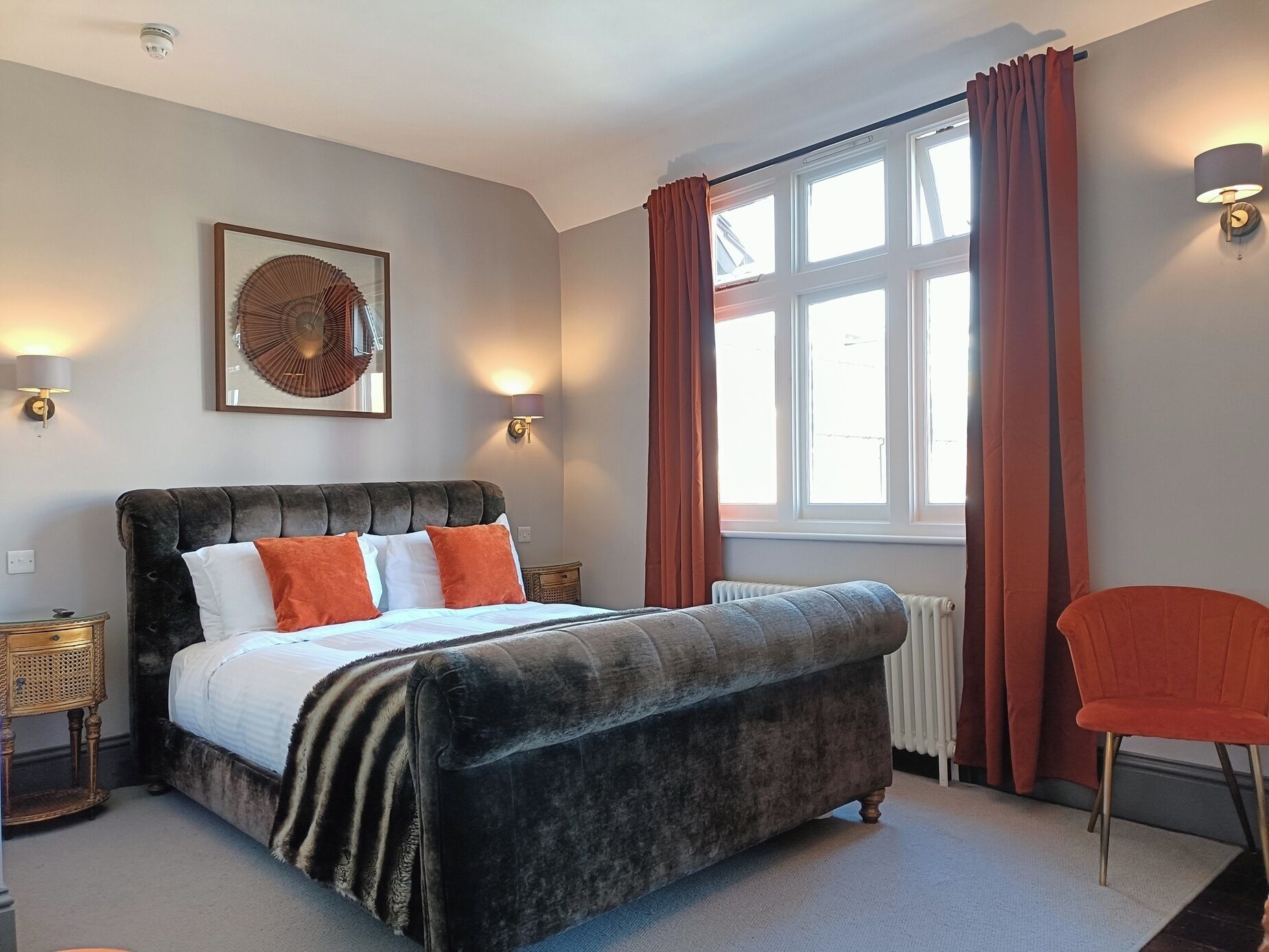 A bedroom with a large bed, a chair, a window with curtains, and wall-mounted lamps.