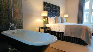 Quarto de hotel moderno com uma banheira grande, cama bem arrumada com cabeceira estampada e pequena mesa lateral com abajur e um vaso de flores.