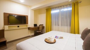 A hotel room with a bed, a desk with a chair, a television mounted on the wall, and a window with yellow curtains.
