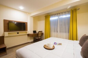 Una camera d'albergo con un letto, una scrivania con una sedia, una televisione a parete e una finestra con tende gialle.