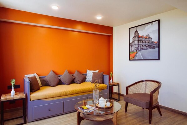 Una habitación con un sofá cama frente a una pared naranja, una silla, una mesita con tetera y un cuadro enmarcado en la pared.