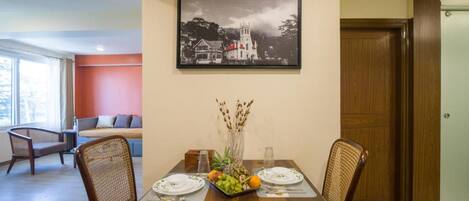 A dining area with a wooden table, two chairs, a fruit bowl, and a framed picture on the wall.