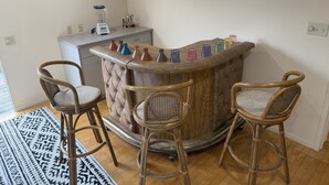 Una zona de bar con mostrador de madera, tres taburetes de mimbre y una variedad de vasos de vidrio de colores.