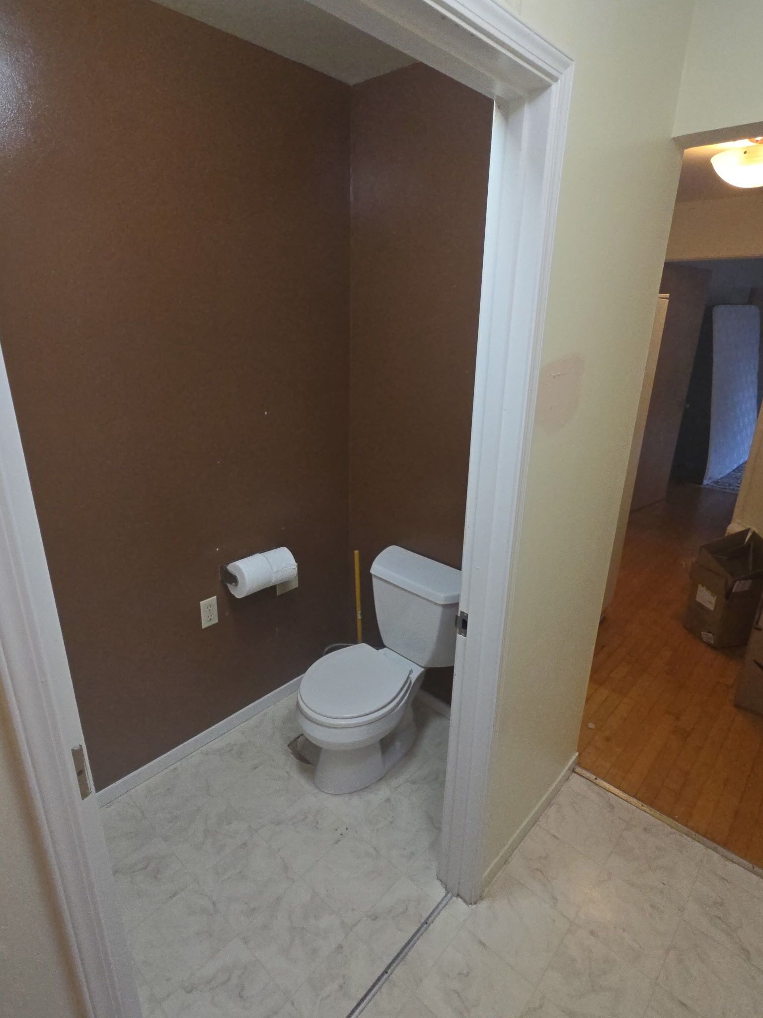 Une petite salle de bain étroite, avec des toilettes, un rouleau de papier toilette et un balai appuyé contre le mur.