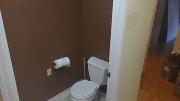 Une petite salle de bain étroite, avec des toilettes, un rouleau de papier toilette et un balai appuyé contre le mur.