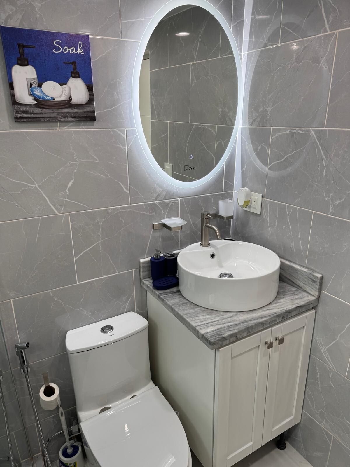 Un baño con inodoro blanco, lavamanos con encimera de mármol y espejo con luz.