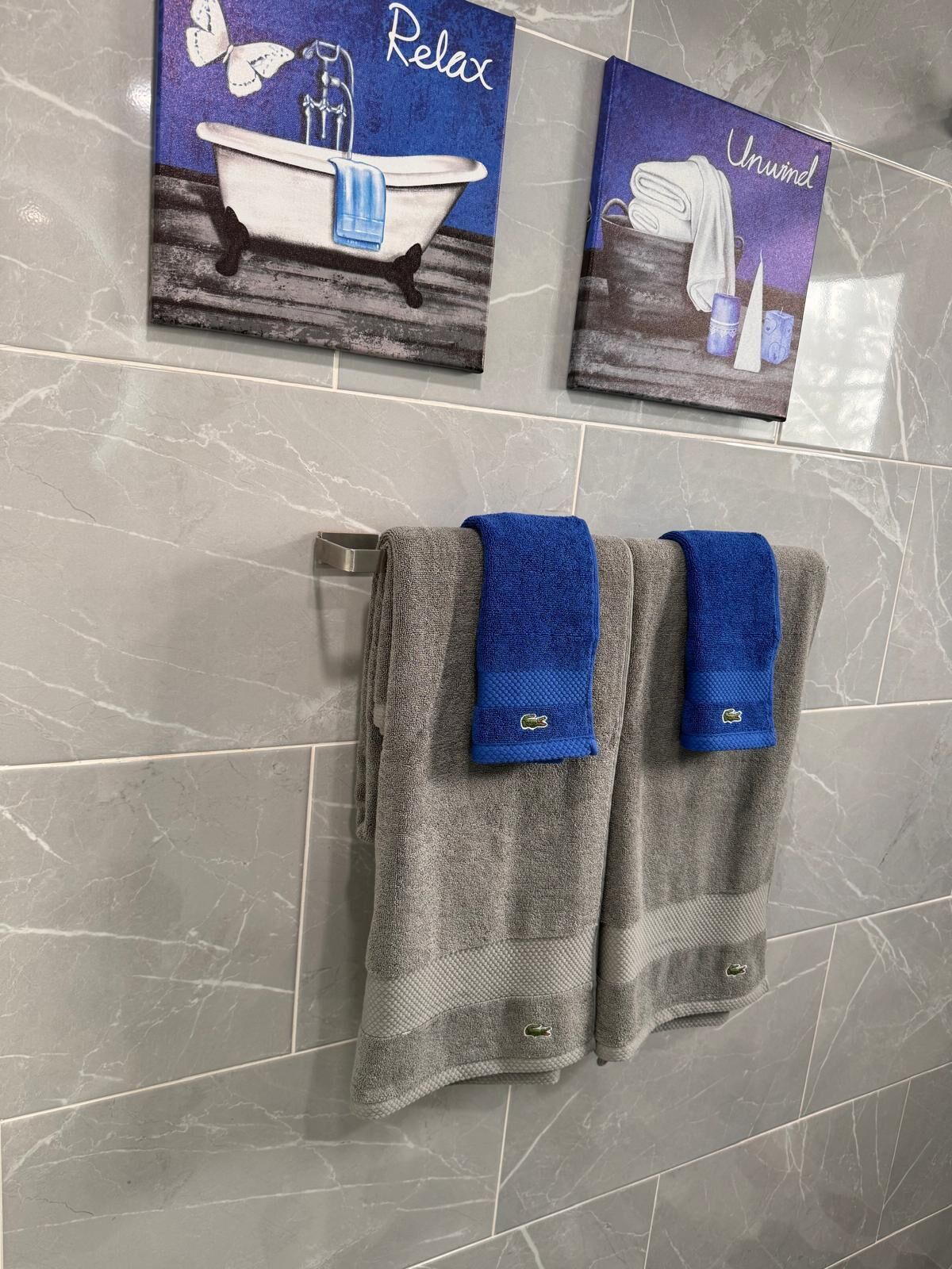 A bathroom with grey marble tiles, two towels hanging on a rack, and two wall art pieces depicting a bathtub and a serene landscape.
