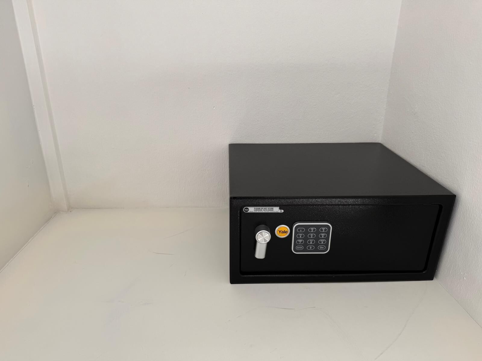 A black safe with a combination dial and a keyhole, placed against a white wall in a corner.