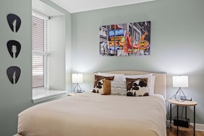 A bedroom with a large bed, two bedside tables, a window with blinds, and a wall art piece.