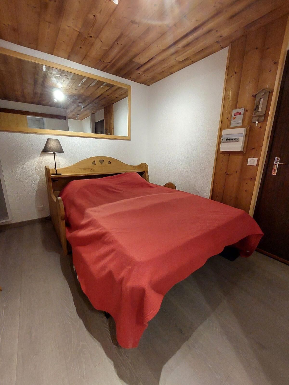 A bedroom with a wooden bed, a red bedspread, a wooden headboard, a nightstand, a lamp, a mirror, and a door with a lock.