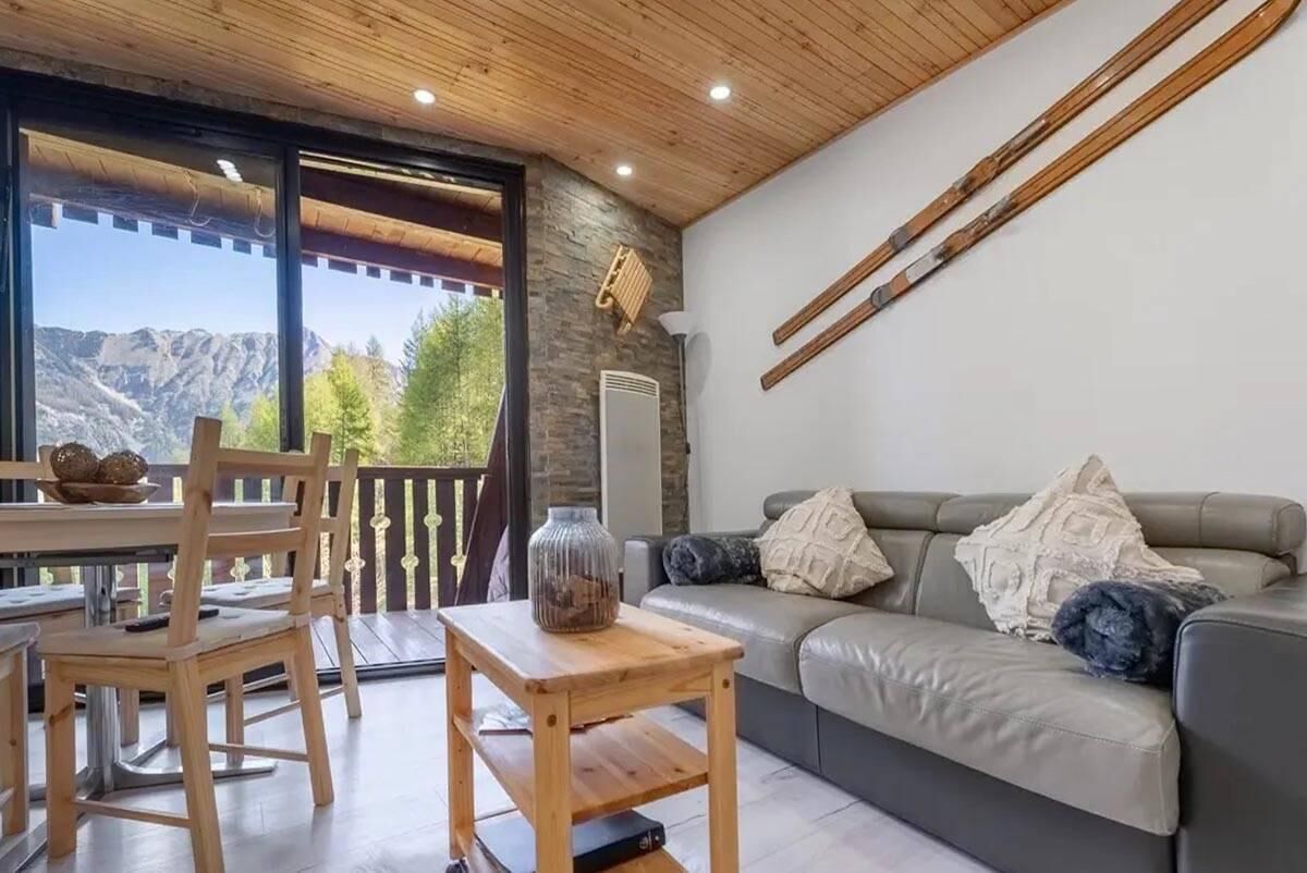 Ein gemütlicher Wohnbereich mit Holzboden, einem grauen Sofa mit Kissen, einem Holztisch mit einer Vase und Bergblick durch große Fenster.