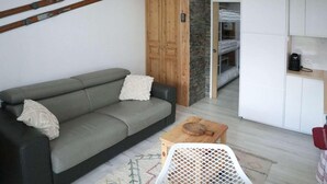 Un espace de vie compact comprenant un canapé, une petite table, un meuble en bois et, au fond, un lit superposé.