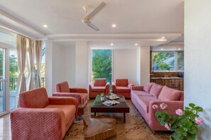 A spacious living room with a large window, a ceiling fan, and a coffee table with a magazine. - Alaya Stays By Elivaas Livelife - Kasauli (Kasauli)