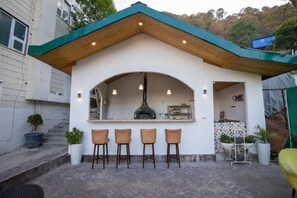 Un pequeño espacio al aire libre con barra, techo de madera, cuatro taburetes y un horno de pizza.
