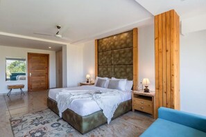 Un dormitorio con una cama grande, cabecera de madera, mesitas de noche con lámparas, un banco y vistas a través de una puerta abierta.