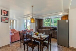 A spacious dining area with a wooden table set for four, surrounded by chairs. A modern pendant light hangs above, and the room features large windows offering a view of greenery outside.