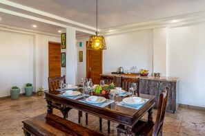Un coin repas avec une table en bois dressée pour six personnes, un luminaire suspendu et un espace cuisine doté d’un plan de travail et de placards.