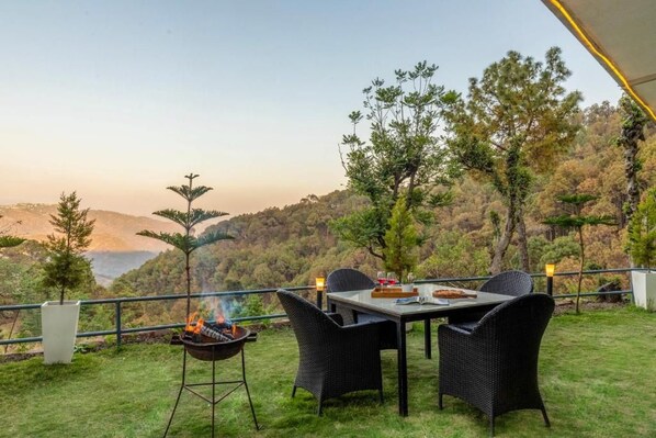 Un área de comedor al aire libre con una mesa y sillas, una parrilla con llamas visibles, y una vista panorámica de un paisaje montañoso.