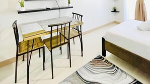Une pièce moderne et bien éclairée, dotée d’un téléviseur à écran plat fixé au mur, d’une table à manger avec des chaises, d’un lit dont la literie est soigneusement faite, et d’un tapis à motifs.