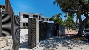 En modern byggnad med en grind vid ingången, en stenmur och fri utsikt över ett bostadsområde med träd och en parkerad bil.