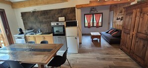 A compact kitchen with a refrigerator, oven, and microwave. There is a dining table with chairs and a sofa in the living area.