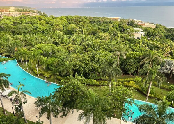 A resort with a large pool surrounded by lush greenery and palm trees, overlooking a coastal area.