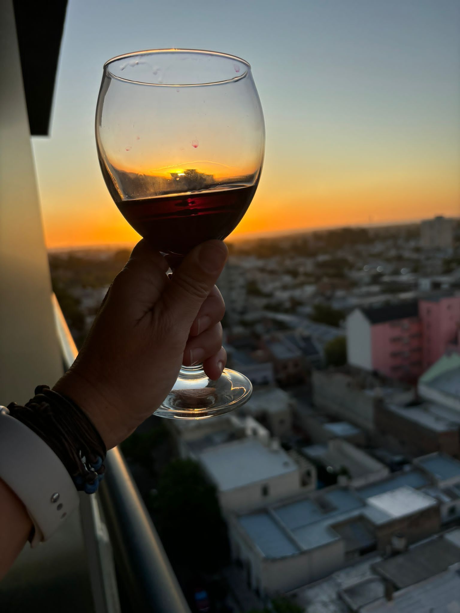 Une main tenant un verre à vin, avec en arrière-plan un coucher de soleil sur une ville.