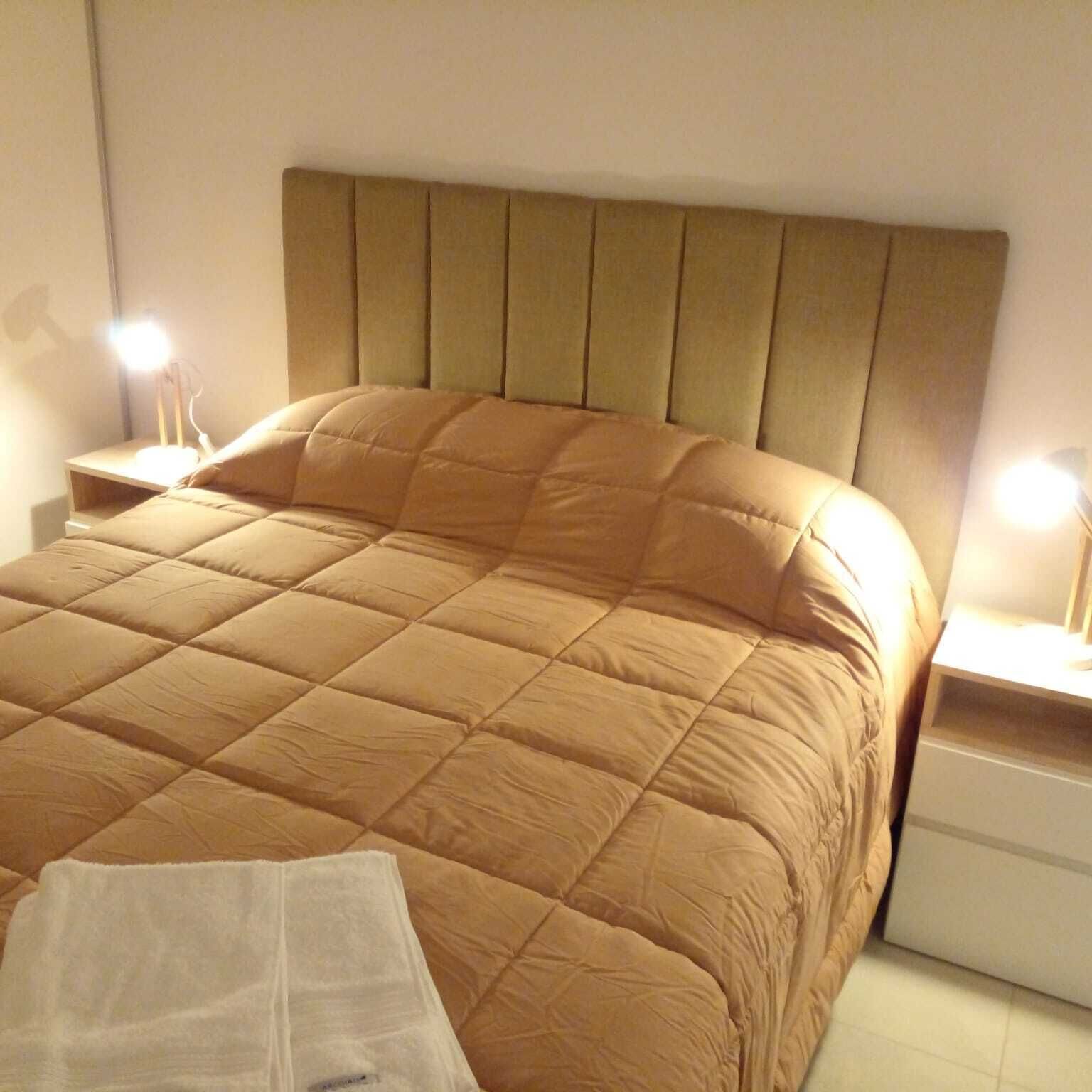 Una cama bien hecha con cabecera, dos mesitas de noche con lámparas y un mueble cajonero blanco.