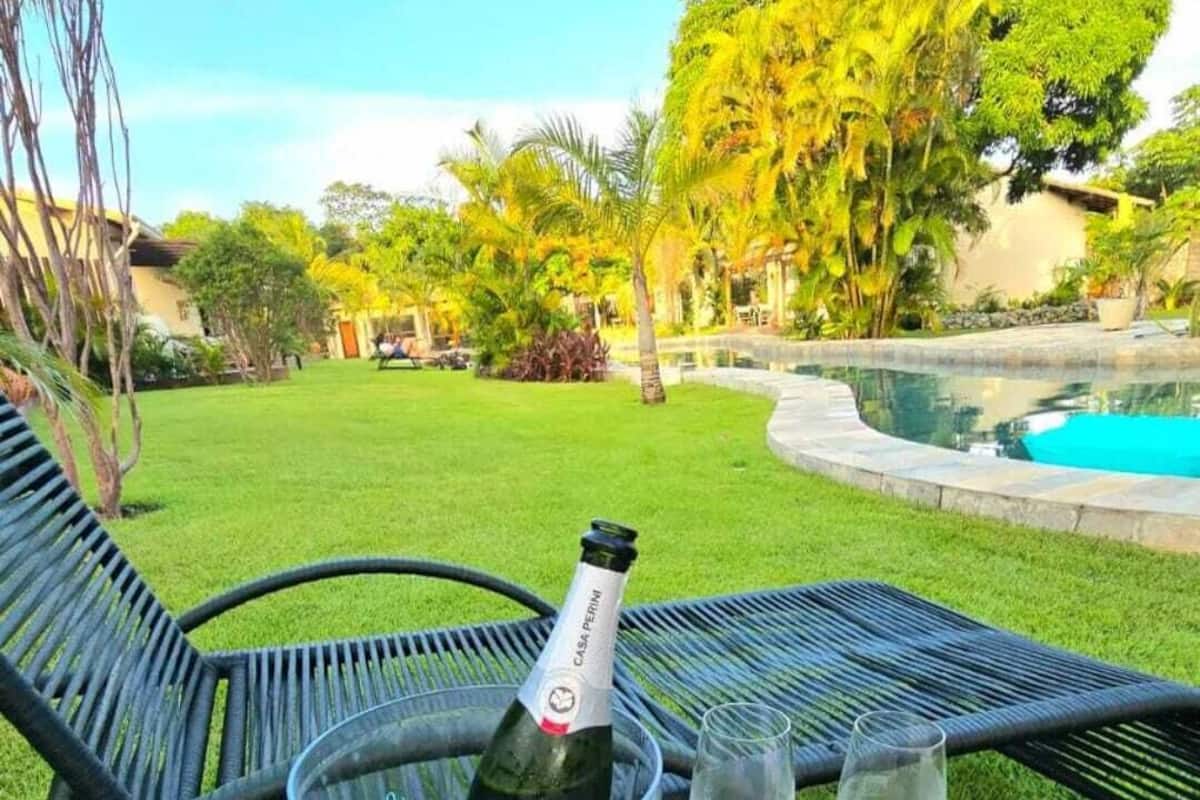 Una mesa de vidrio con una botella de champán y dos copas, ubicada en un jardín exuberante con una piscina al fondo.