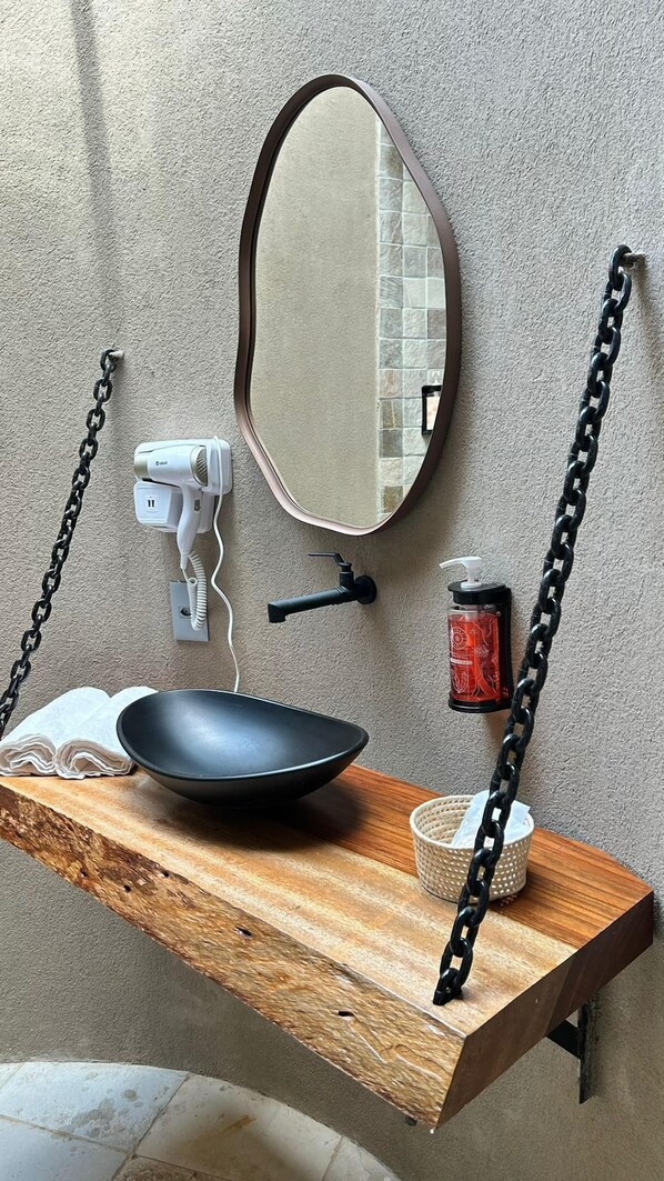 Un baño con tocador de madera, un lavabo negro, un espejo redondo, un dispensador de jabón y un perchero para toallas.