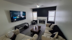 A living room with a flat-screen TV mounted on the wall, a fireplace, two sofas, a coffee table, and a ceiling fan. - Modern Family Retreat w/ Office & Fast WiFi (East Hartford)