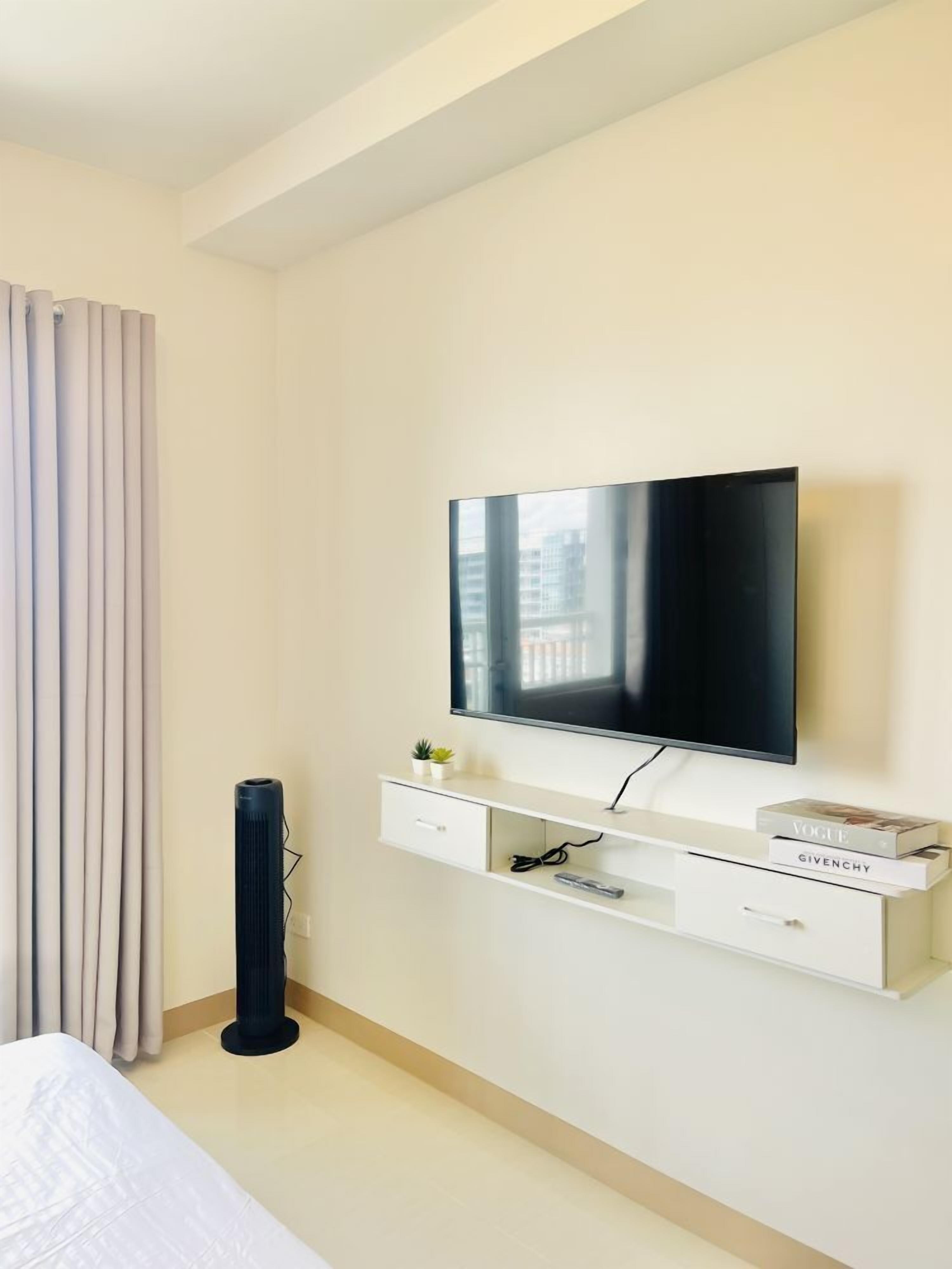 A modern hotel room with a flat-screen TV mounted on a white console, a bed with a white headboard, a black floor lamp, and a window with curtains.