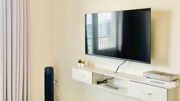 A modern hotel room with a flat-screen TV mounted on a white console, a bed with a white headboard, a black floor lamp, and a window with curtains.