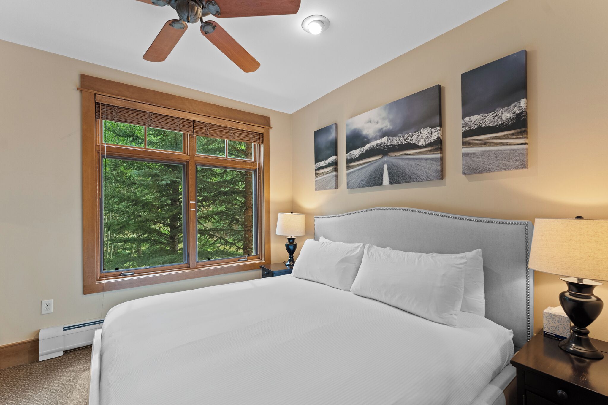A hotel room with a large bed, a ceiling fan, a window with blinds, and three landscape paintings on the wall.