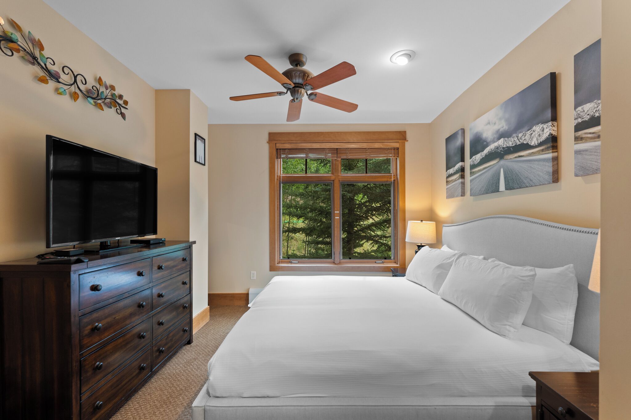 Une chambre avec un lit, un téléviseur posé sur une commode, un ventilateur de plafond, une fenêtre donnant sur des arbres et des œuvres d’art accrochées aux murs.