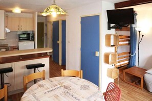 A compact living space with a kitchenette, a dining area with a patterned tablecloth, a television mounted on the wall, and a red chair.