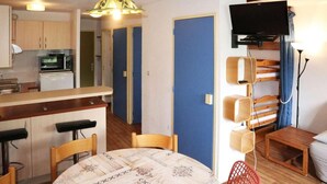 Un espace de vie compact comprenant un coin cuisine, un coin repas avec une nappe à motifs, un téléviseur fixé au mur et un petit coin salon avec un banc en bois et une chaise rouge.