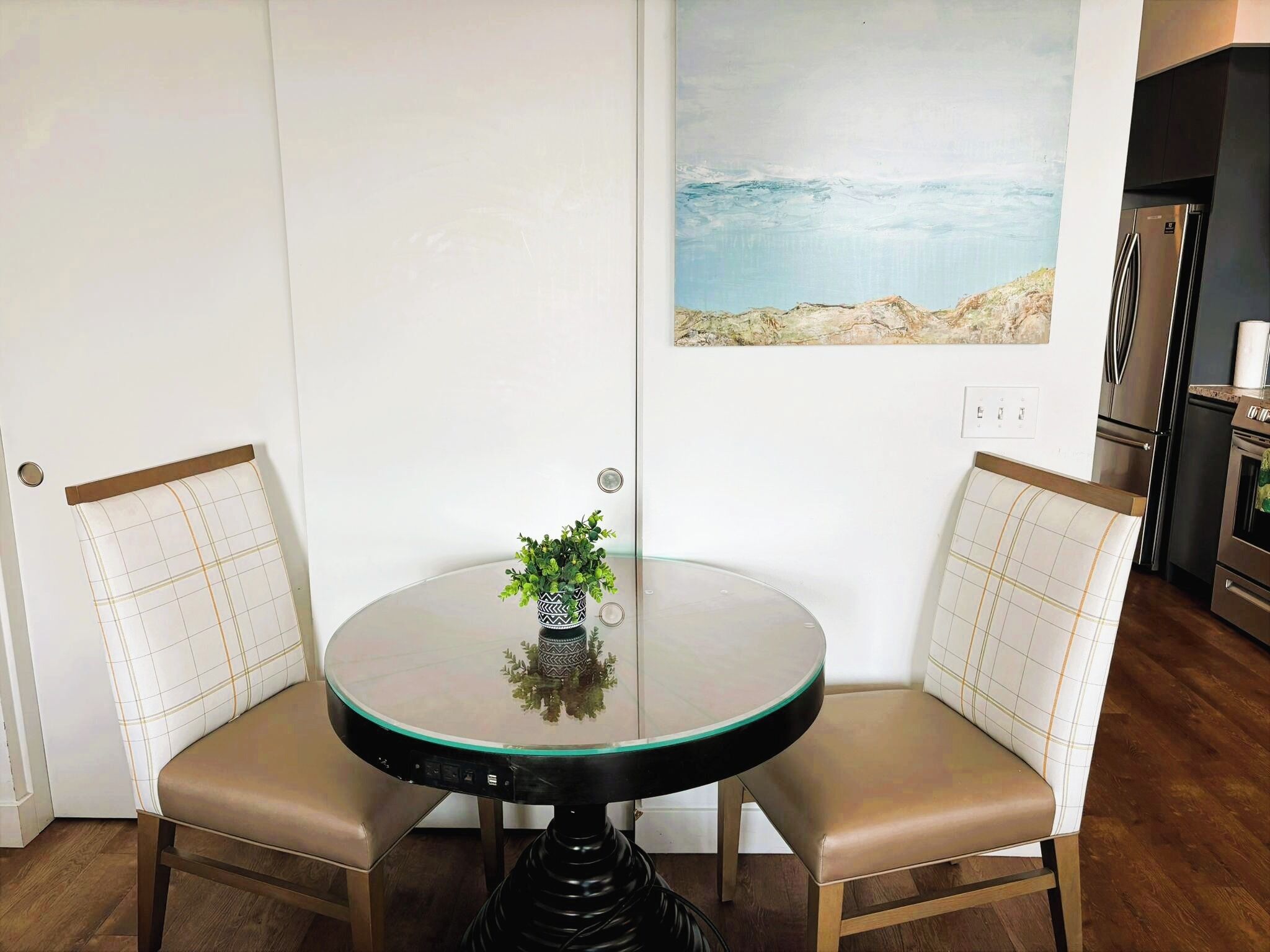 A dining area with a round glass table, two cushioned chairs, a small potted plant, and a painting of a beach scene on the wall.