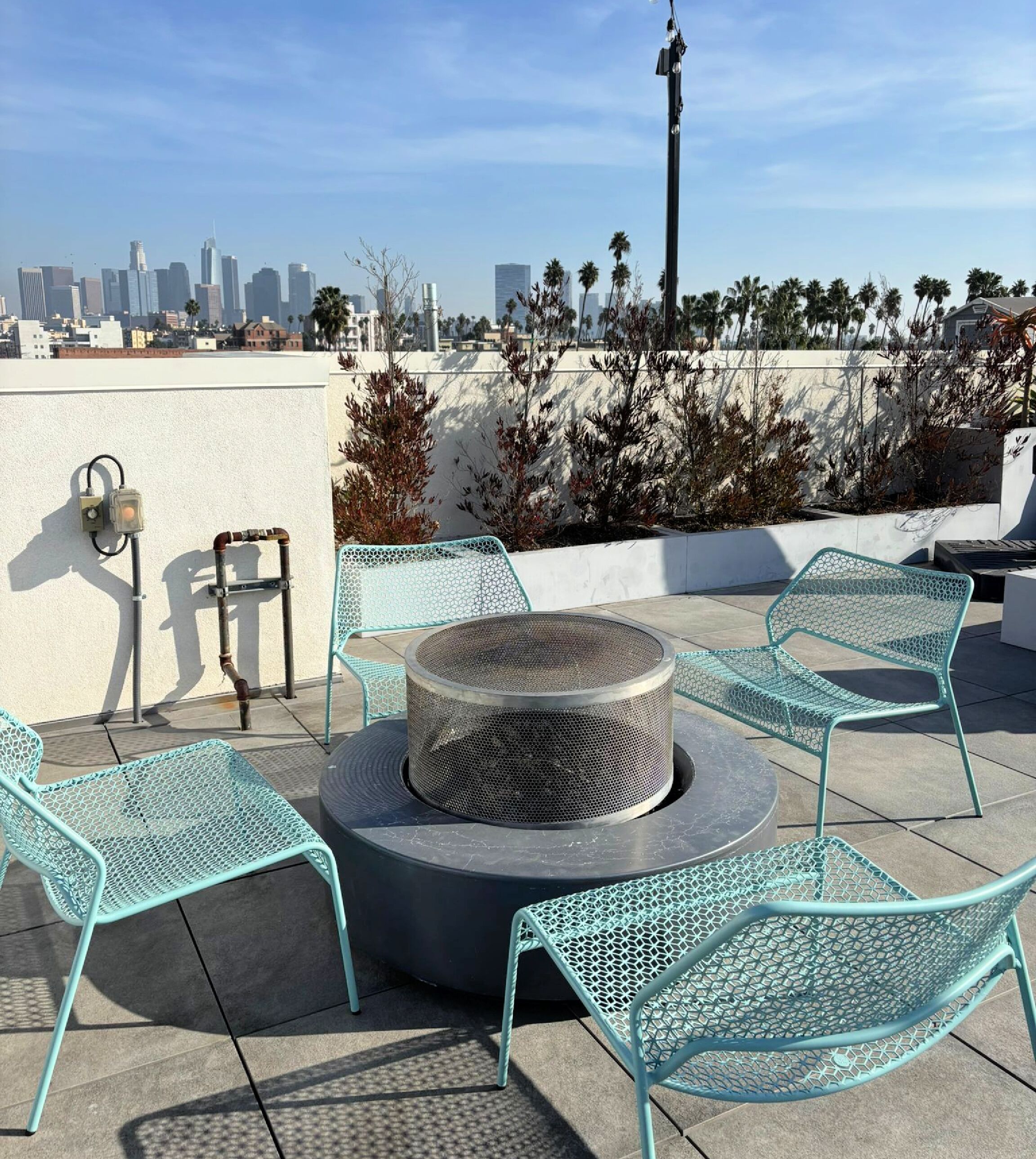 A rooftop patio with a round fire pit, metal chairs, and a city skyline view.